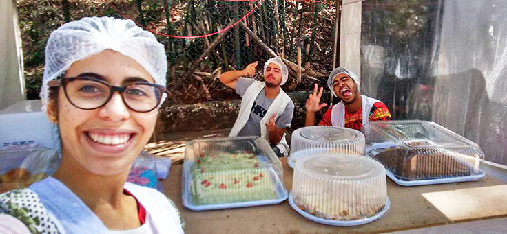 Imagem mostra Pamela sorridente em primeiro plano. Atrás, duas pessoas sorriem também. Elas estão atrás de uma mesa com bolos