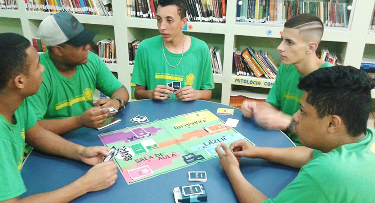 Jovens sentados em mesa com tabuleiro jogam o jogo Se Vira do Pense Grande