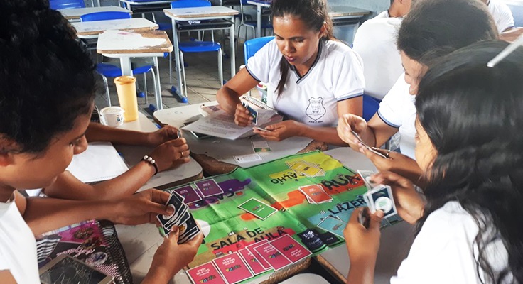 Jovens sentados em mesa com tabuleiro jogam o jogo Se Vira do Pense Grande