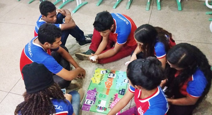 Jovens sentados em mesa com tabuleiro jogam o jogo Se Vira do Pense Grande