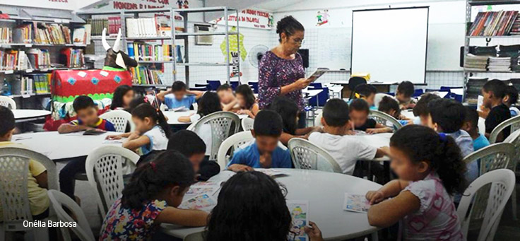 Professora circula em sala de aula com crianças sentadas em mesas circulares.
