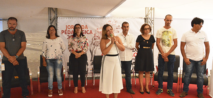 Grupo de pessoas em pé em frente a um banner onde se lê “Jornada Pedagógica”. Uma moça de macacão branco está falando ao microfone.