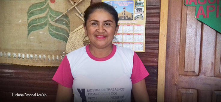 Em imagem horizontal, professora indígena Luciana Pascoal Araújo, de camiseta branca e rosa no ambiente escolar