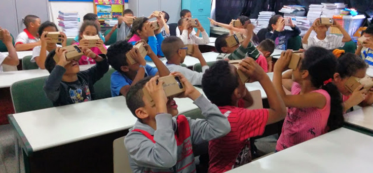 Crianças em sala de aula sentadas em suas carteiras usam Google Cardboard, óculos de realidade virtual com estrutura de papel craft e baixo custo.