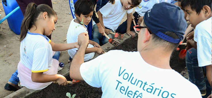 Voluntário aparece ao lado de crianças em ação de manutenção da horta da Escola Municipal Lili Benchimol