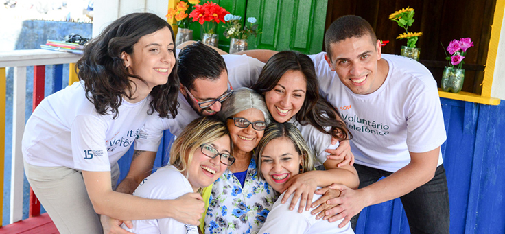 Voluntários do Vacaciones Solidárias rodeiam e abraçam educadora em ação na sala de leitura da Escola Municipal Lili Benchimol