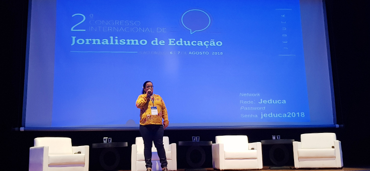 Educadora Maria do Socorro está em pé no palco do Jeduca. Ela usa blusa amarela e óculos. Está falando ao microfone sobre a vivência em escola pública
