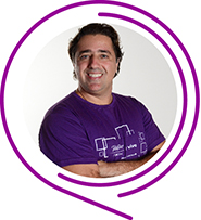 Na imagem, o voluntário Alexandre de Souza Pinto posa de braços cruzados e usando camiseta púrpura do Programa de Voluntariado. Ele tem cabelos pretos e sorri para a foto