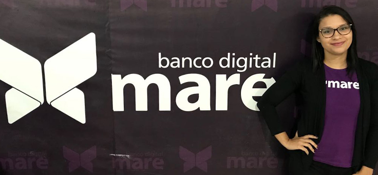 Mulher usando camiseta do Banco Digital Maré posa à frente do logotipo da startup. Ela usa óculos, veste blazer preto e está sorrindo para a foto.