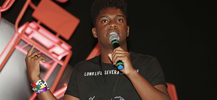 Aluno Luã dos Santos Silva sua camiseta preta e fala ao microfone no palco do Educação 360