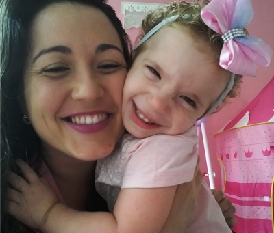 Paloma Almeida em cabelos compridos e usa batom rosa. Ela está abraçada à filha, Giovanna, que tem 3 anos e usa um laço rosa na cabeça. As duas sorriem para a foto