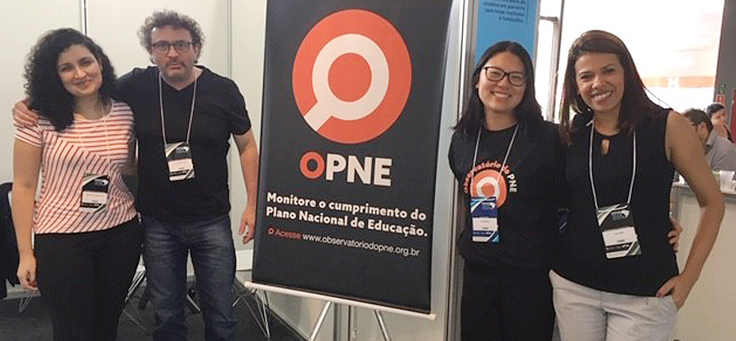 Na imagem, quatro pessoas, três mulheres e um homem posam ao lado de banner com logo da plataforma do OPNE, que monitora a educação brasileira.