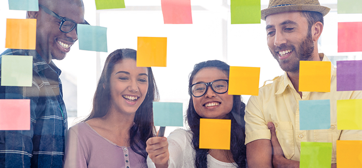 Imagem ilustra a Semana Global do Empreendedorismo e mostra grupo de quatro jovens olhando para post its colados em uma parede transparente