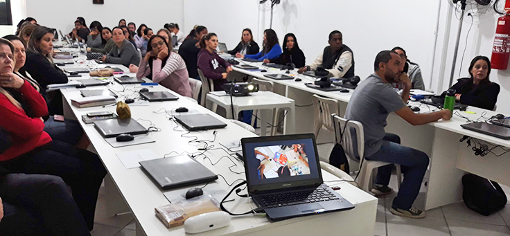 Professores estão sentados em frente a computadores, assistindo a projeção durante formação continuada da Fundação Telefônica Vivo em parceria com a Undime.