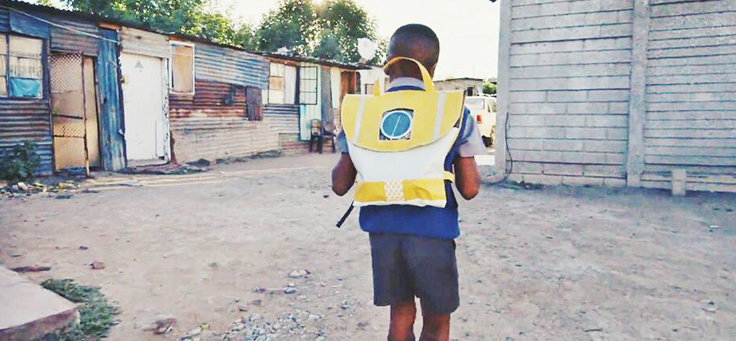 Na imagem menino está usando a bolsa do projeto Repurpose Schoolbags, que auxilia na educação de crianças, enquanto anda por rua sem asfalto.
