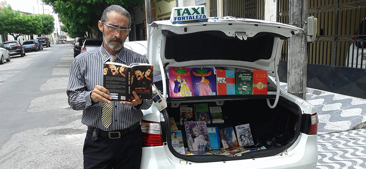 Motorista conhecido como Carlos Careca está com um livro na mão posando em frente ao táxi que dirige – matéria sobre o bibliotaxi foi destaque em empreendedorismo social em 2018.