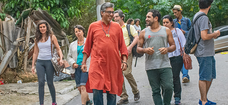 Manish Jain anda pelo Jardim Ângela em São Paulo. Ele está usando uma bata laranja e está rodeado por jovens do bairro.