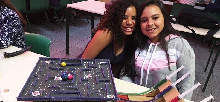 Duas alunas estão posando em frente a tabuleiro de jogo feito com sucata em aula sobre robótica da professora Débora Garofalo, finalista do Nobel da Educação.
