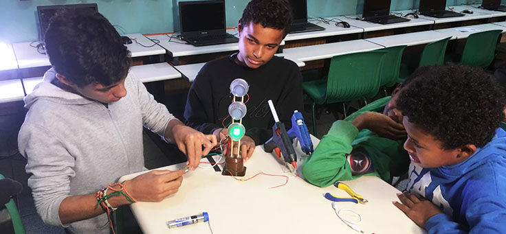 Três alunos estão segurando dispositivo que emite luz verde em aula de robótica da professora Débora Garofalo, finalista do Nobel da Educação.