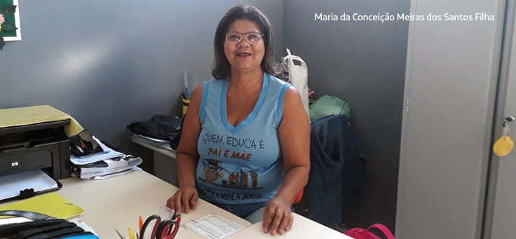 Gestora Conceição sorri para a câmera sentada em sua mesa. Ela usa blusa azul onde se lê: “Quem educa é pai e mãe”.
