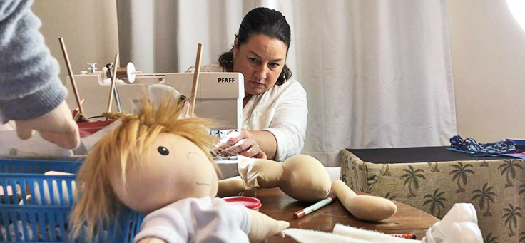Amy Jandrisevits aparece na imagem costurando em uma máquina de costuras. Uma boneca está em primeiro plano