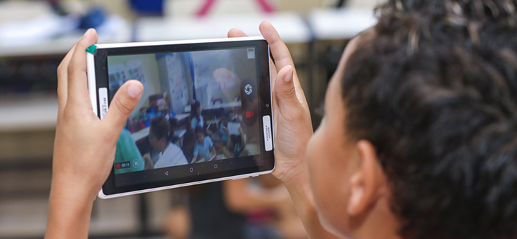 Aluno segura tablet na altura dos olhos para tirar foto da sala de aula durante evento de entrega de kit tecnológico do Aula Digital, que incentiva a inclusão digital, em Viamão (RS).