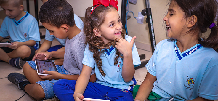 Duas meninas se olham e sorriem enquanto usam tablets distribuídos durante evento de entrega de kits tecnológicos do Aula Digital, programa que incentiva a inclusão digital, em Viamão (RS).