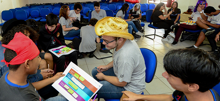 Estudantes usando chapéus estão sentados frente a frente olhando para um canvas impresso em um papel sulfite. A metodologia do Pense Grande ensina atividades sobre cultura empreendedora.