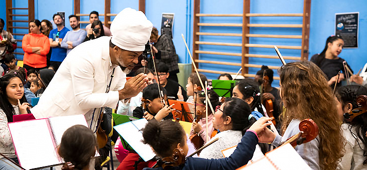 Carlinhos Brown, que usa turbante e roupas brancas, está fazendo reverência a um grupo de meninos e meninas da Fundación para la Acción Social por la Música, enquanto tocam violino em festival realizado em colégio em Madri.
