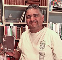 Paulo César Marciano, o PC, idealizador da Editora Gráfica Heliópolis, está segurando o primeiro livro publicado dele, Melissa, com uma estante com livros ao fundo.