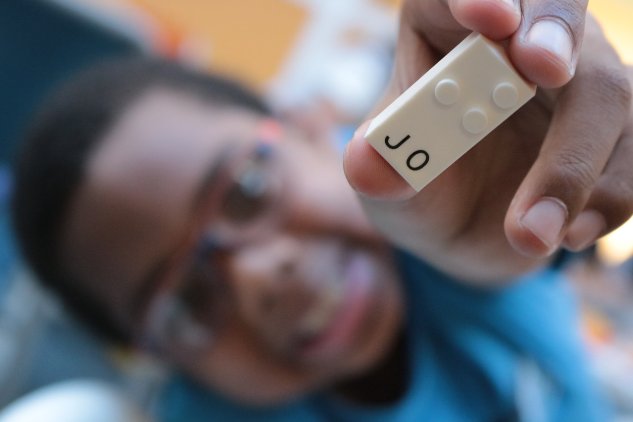 Em primeiro plano a mão de um menino segura uma peça branca com pontos em alto relevo, que faz parte do projeto de educação inclusiva da Lego. Embaixo dos pontos se lê as letras J e O. Ao fundo, desfocado está o rosto do garoto, que usa óculos e sorri.