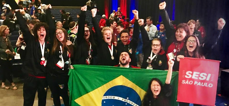 Na imagem, diversos jovens brasileiros posam para foto durante campeonato de robótica. Eles estão com os braços levantados e seguram uma bandeira do Brasil e outra do SESI São Paulo.