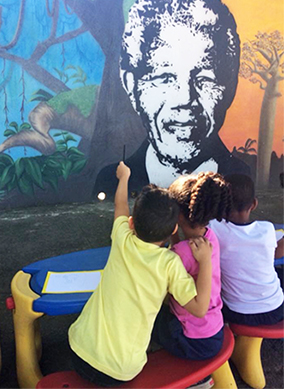 Três alunos estão sentados em frente a muro da EMEI Nelson Mandela, onde está desenhado o rosto de Madiba com árvores típicas da savana africana ao fundo.