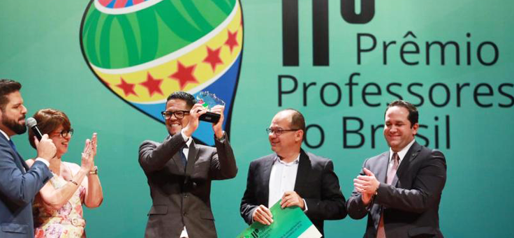 Professor Isaías Silva, vestindo terno e óculos, está erguendo troféu no palco do 11º Prêmio Professores do Brasil.