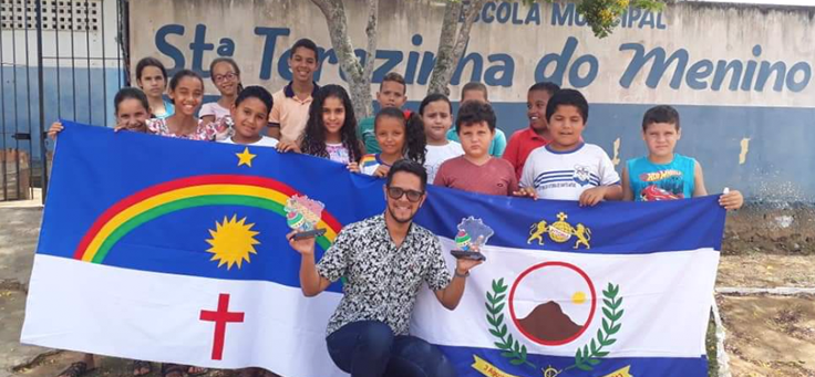 Educador Isaías Silva está agachado, segurando troféus do Prêmio Professores do Brasil. Atrás dele está um grupo de alunos, mostrando as bandeiras de Pernambuco e de Vitória de Santo Antão.