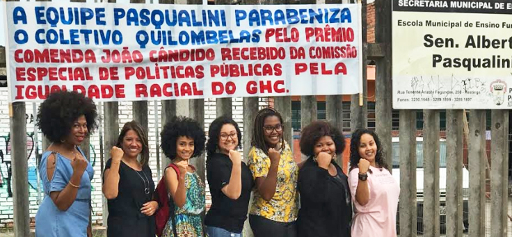 Mulheres do coletivo feminista Quilombelas estão posando com os punhos levantados indicando força. Acima delas há uma faixa de homenagem por um prêmio conquistado.