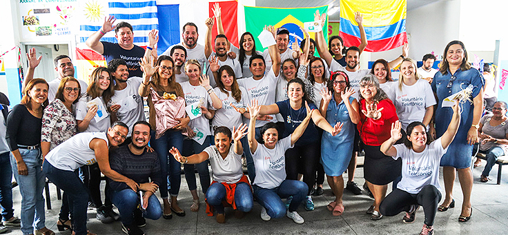 Grupo de 15 colaboradores do programa de voluntariado Vacaciones Solidárias posa na Escola Municipal de Ensino Fundamental Frei Fernando com bandeiras de seus países ao fundo.