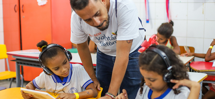 Colaborador do programa de voluntariado Vacaciones Solidárias está observando duas alunas da Escola Municipal de Ensino Fundamental Frei Fernando que estão sentadas na carteira escrevendo.