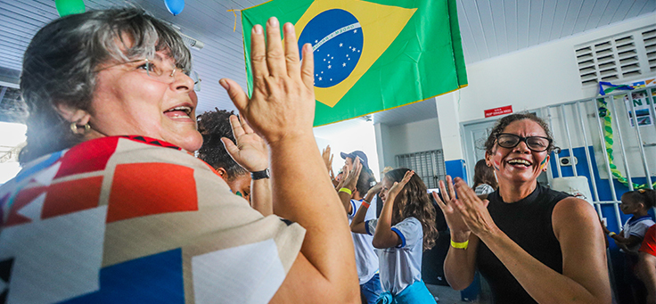 Duas colaboradoras do programa de voluntariado Vacaciones Solidárias batem palma, com bandeira do Brasil ao fundo.