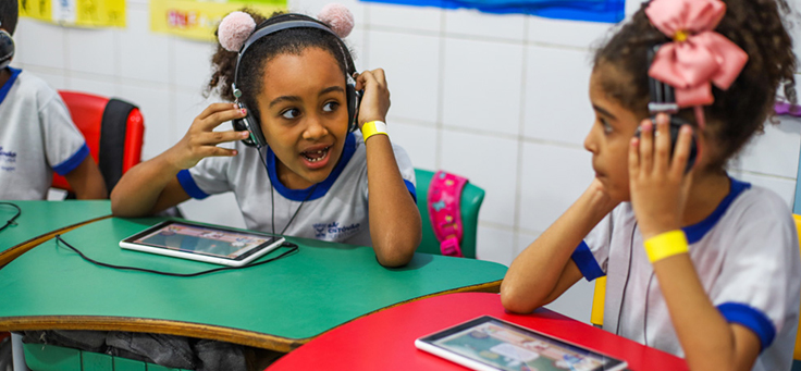 Duas crianças estão sentadas em carteira com fones no ouvido em sala da Escola Municipal de Ensino Fundamental Frei Fernando, que recebeu o programa de voluntariado Vacaciones Solidárias.