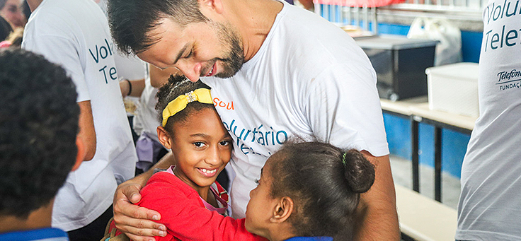 Colaborador está abraçando duas alunas da Escola Municipal de Ensino Fundamental Frei Fernando, que recebeu o programa de voluntariado Vacaciones Solidárias.