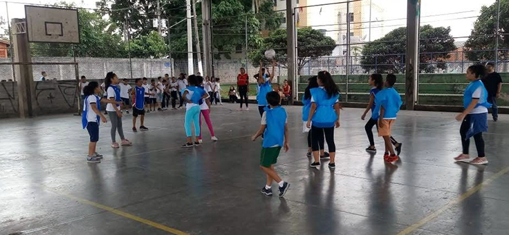 Crianças estão jogando vôlei em quadra usando coletes na cor azul claro durante aula de Educação Física, disciplina é valorizada pelo conceito de escola ativa.