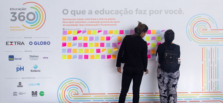 Imagem mostra pessoas colando post-its coloridos em um banner onde se lê “O que a Educação faz por você”. Ao lado se vê o nome do evento: Educação 360 e os logotipos de parceiros e patrocinadores.