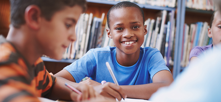 Menino está sentado em meio a livros, sorrindo e segurando um lápis para ilustrar pauta sobre formação de educadores para melhorar a alfabetização.