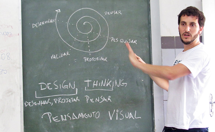 Imagem mostra formador usando uma camiseta do Pense Grande apontando para uma lousa onde se vê um desenho ao fundo e palavras relacionadas à metodologia como design thiking e pensamento visual