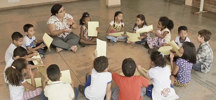 Imagem mostra a professora Elenice sentanda no chão com seus alunos em uma roda.