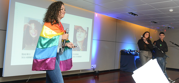 Imagem mostra jovem enrolada em bandeira LGBT. Dois colegas a observam no palco