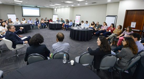 Integrantes da rede do Escola Digital estão sentados em roda conversando com especialistas, em meio a charts e um telão, durante o encontro ocorrido em São Paulo.