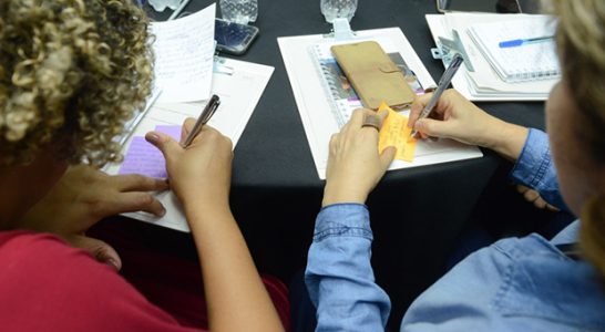 Duas integrantes da rede do Escola Digital aparecem de costas, escrevendo em post its durante encontro ocorrido em São Paulo.