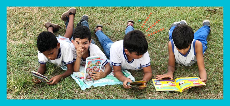 Quatro meninos estão deitados em gramado lendo livros em atividade do projeto de estímulo à leitura piquenique literário, criado por meio do Aula Digital.